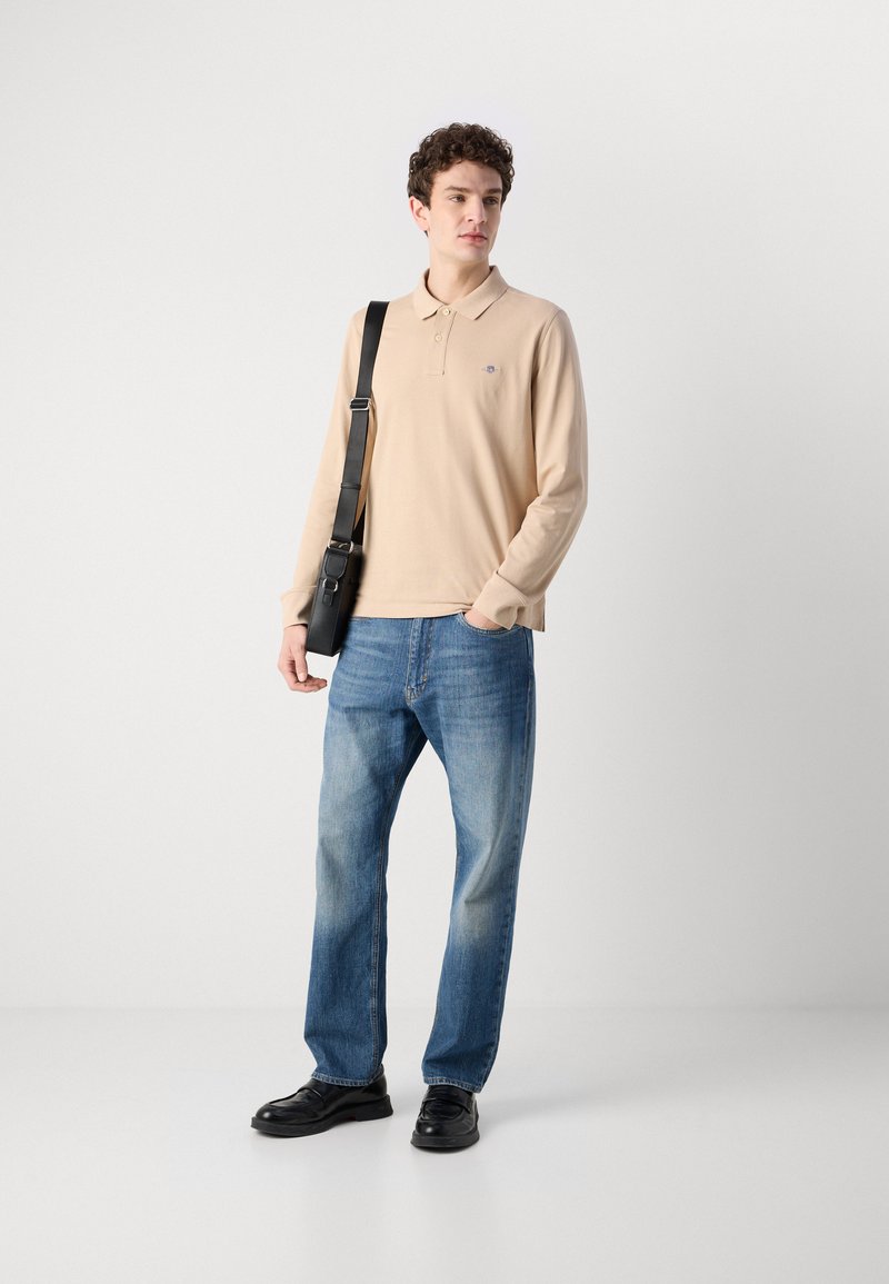 Beige Langarm-Polo mit Kragen und dezentem Logo, kombiniert mit hellblauen geraden Jeans und schwarzen Schnürschuhen.