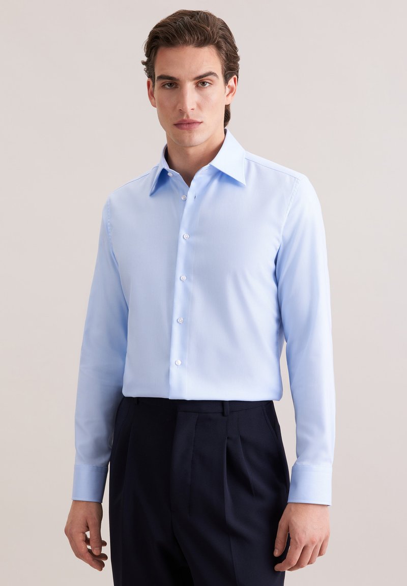 Jeune homme portant une chemise à manches longues bleu clair rentrée dans un pantalon sombre taille haute, debout devant un fond uni.