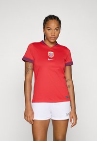 T-shirt rouge à manches courtes, avec un col accentué de bleu et des poignets à motifs, présente un logo central, associé à un short blanc, avec la marque Nike bien visible.