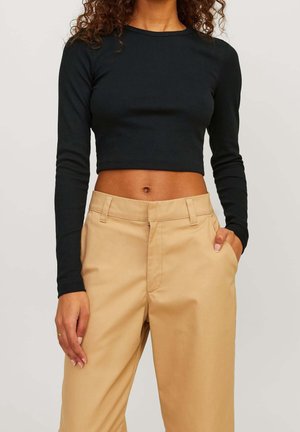 Femme portant un crop top noir à manches longues et un pantalon beige taille haute, les mains dans les poches, debout devant un fond uni.