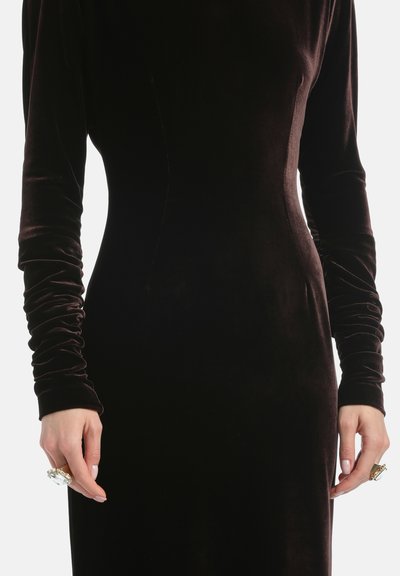 Czarna, aksamitna sukienka wieczorowa z długimi, marszczonymi rękawami. Charakteryzuje się gładką teksturą i dopasowanym krojem. Ręka ozdobiona dużym pierścionkiem.