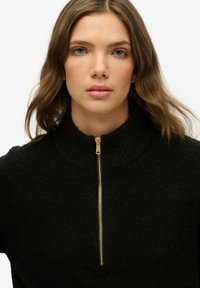 Svart tröja med hög krage, utrustad med en guldfärgad dragkedja. Tyget är strukturerat, mjukt och stickat, vilket ger en mysig och avslappnad design.