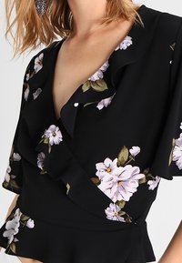 Schwarze florale Wickelbluse mit gerafftem Halsauschnitt, verziert mit helllila Blumen und grünen Blättern, aus strukturiertem Stoff gefertigt. Detail mit Peplum-Saum.
