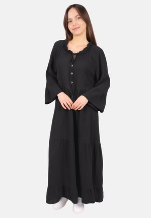 Frau steht, trägt ein langes schwarzes Kleid mit Knöpfen und weiten Ärmeln, kombiniert mit weißen Turnschuhen, vor einem einfarbigen hellen Hintergrund.