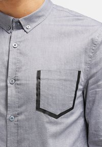 Camisa de algodón gris con cuello abotonado, que presenta un bolsillo con contorno negro. Textura suave y diseño clásico con botones azules.