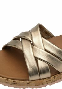 Guldmetallisk slid sandal med korslagda remmar, slät yta och en brun vadderad fotbädd ovanpå en korkliknande sula.