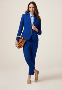 Niebieski garnitur z dopasowaną marynarką i pasującymi spodniami. Biała koszula w niebieskie paski, połączona z brązową torbą na ramię i beżowymi balerinami.