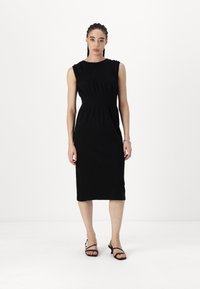 Melna midi kleita ar pieguļošu priekšdaļu, savelku tekstūru jostas daļā un bez piedurknēm. Audums ir mīksts un gluds. Valkāta ar melnām sandālēm.