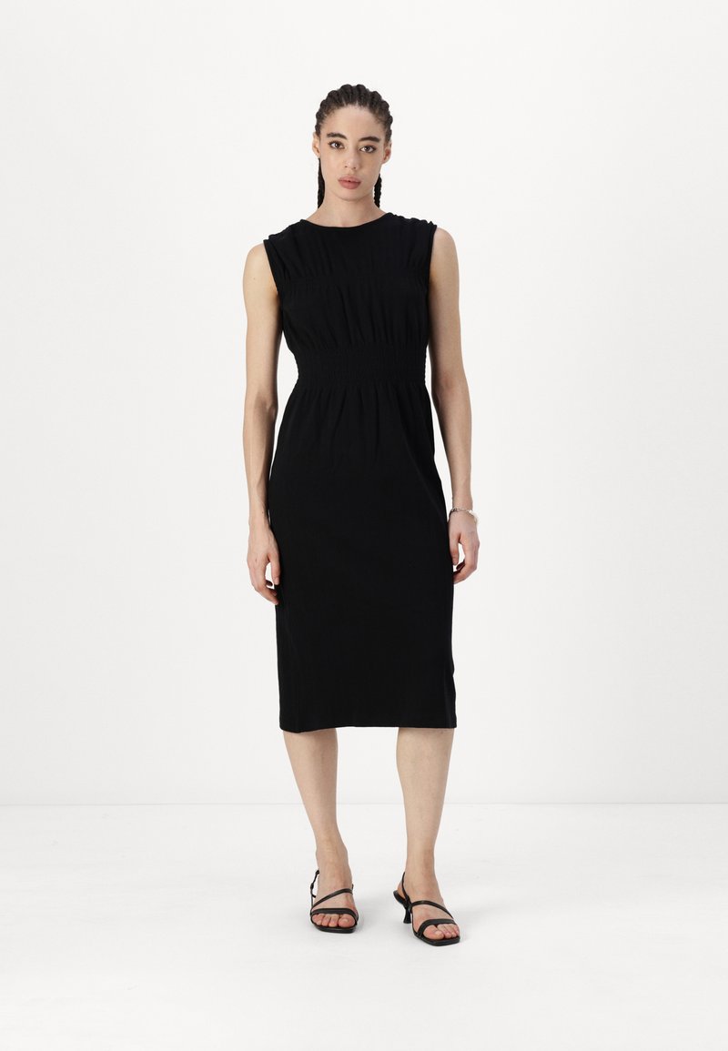 Melna midi kleita ar pieguļošu priekšdaļu, savelku tekstūru jostas daļā un bez piedurknēm. Audums ir mīksts un gluds. Valkāta ar melnām sandālēm.