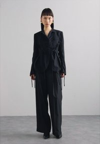 Schwarze taillierte Jacke mit Kordelzugakzenten, kombiniert mit weiten schwarzen Hosen. Glatte Stoffe, strukturierte Form und minimalistisches Design.