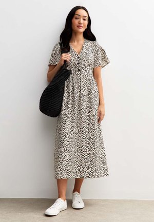 Robe maxi en crème avec des pois noirs, manches courtes évasées, encolure en V et taille cintrée. Associée à un sac noir texturé et des baskets blanches.