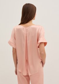 Blouse en satin rose clair avec des manches courtes et un col rond, dotée d'une fente au dos et d'une coupe décontractée. Texture lisse et douce.