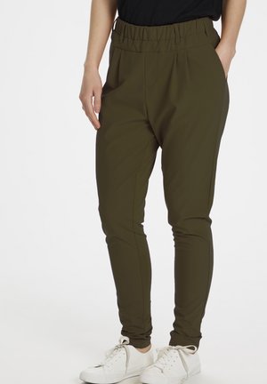 Osoba ubrana w oliwkowo-zielone spodnie zwężane z elastycznym pasem oraz białe trampki, stojąca z jedną ręką w kieszeni.