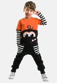 Oranges und schwarzes Outfit mit gestreiften langen Ärmeln, das eine verspielte Monstergrafik zeigt. Schwarze Hose mit gestreiften Taschen und Turnschuhen.