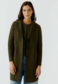 Manteau en laine verte avec un col cranté, des boutons à l'avant et des manches longues, superposé sur un pull en laine côtelé vert et un jean bleu.