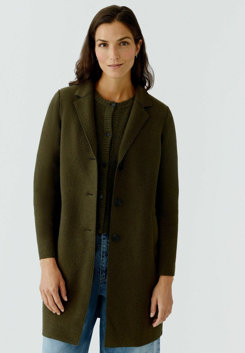 Manteau en laine verte avec un col cranté, des boutons à l'avant et des manches longues, superposé sur un pull en laine côtelé vert et un jean bleu.