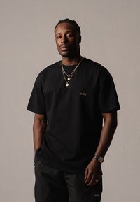 Black cotton t-shirt with short sleeves, featuring a small red logo on the left chest. Worn with black shorts and layered gold necklaces.