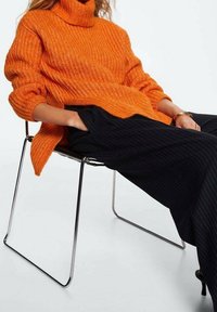 Personne assise sur une chaise en métal portant un pull à col roulé orange oversized en maille et un pantalon large à rayures noires.
