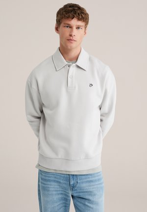 Jonge man staand met handen achter de rug, gekleed in een lichtgrijs poloshirt met lange mouwen over een gedempt groen T-shirt en blauwe spijkerbroek.