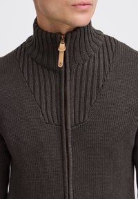 Maglione grigio scuro a maglia con colletto alto, design a coste e una cerniera visibile con un tirazip in pelle beige. Texture liscia in tutto il capo.