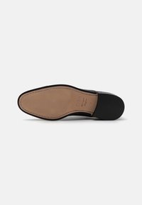 Suela de zapato de cuero negro con una forma redondeada y suave, que presenta una parte inferior de goma color tostado y un diseño texturizado sutil. Acentos de costura visibles.