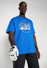 Zila kokvilnas t-krekls ar baltu "Nike Golf" grafiku un apļa rakstu. Īsas piedurknes, brīvs piegriezums, valkāts ar baltu golfa cimdiņu un melniem šortiem.