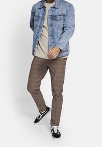 Ljusblå denimjacka med broderi "Impulsive", bärs över en beige skjorta. Bruna rutiga byxor och svarta sneakers fulländar stilen.