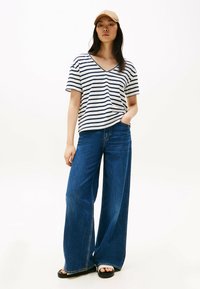 Camiseta de rayas en v en blanco y azul marino, combinada con unos jeans de pierna ancha en azul. El modelo lleva una gorra beige clara y sandalias negras, de pie con confianza.