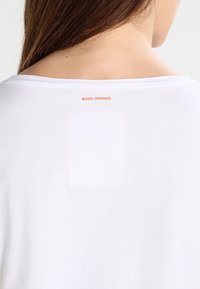 Bílá tričko s jemnou texturou, na zadní straně s obdélníkovou bílou visačkou a nápisem "BOSS ORANGE" tištěným světle oranžovou barvou nad ní.