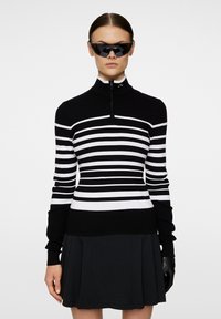 Randig svartvit stickad tröja med hög krage, dragkedja och åtsittande design. Bärs med en svart plisserad kjol och svarta solglasögon.