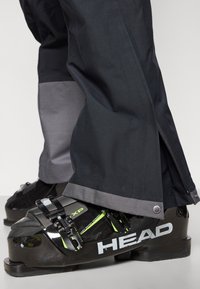 Crne skijaške čizme sa zelenim naglascima i podesivim kopčama, uparene s tamnim hlačama koje imaju sive rubove i detalj bočnog zatvarača.