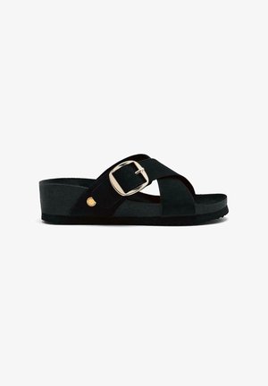 Sandalia de ante negra con diseño de tiras cruzadas, hebilla ajustable y suela plana y texturizada. Presenta un acento dorado redondo en el lado exterior.