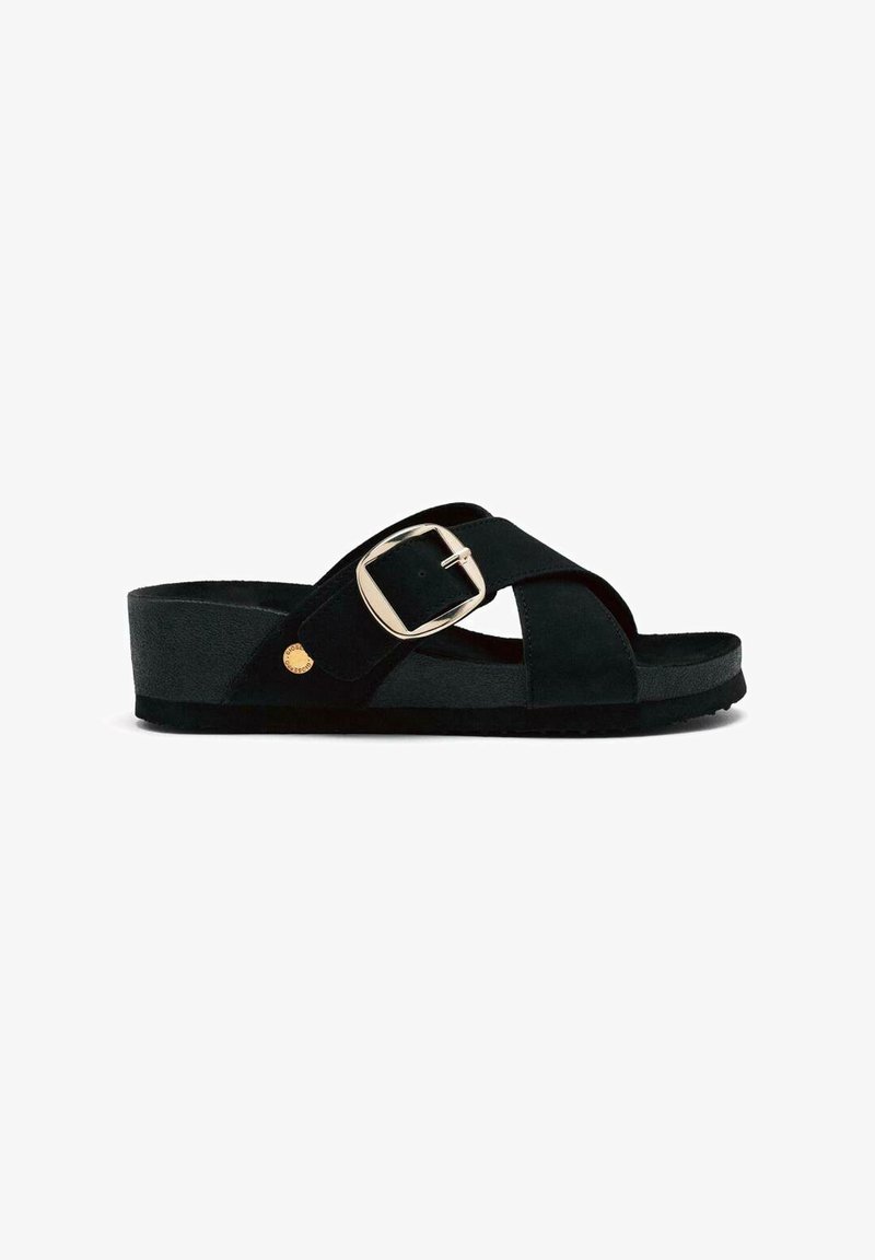 Μαύρη σουέτ σανδάλια με σχέδιο διασταυρωμένων ιμάντων, ρυθμιζόμενο κούμπωμα και επίπεδη, υφή σόλα. Περιλαμβάνει μια στρογγυλή χρυσή λεπτομέρεια στην εξωτερική πλευρά.