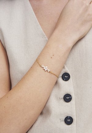 Guld kædearmbånd med tre små blomstvedhæng båret på et håndled over en beige ærmeløs knaplukket beklædning.