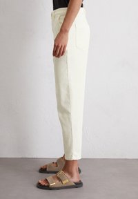 Pantalones chinos color crema con corte recto, que cuentan con bolsillos laterales y una textura de tela suave. Combinados con sandalias marrones y una blusa negra.