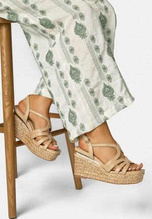 Voeten met beige sandalen met bandjes en vlechtwerk op de hakken, gekruist bij de enkels, rustend op een houten stoelbeen.