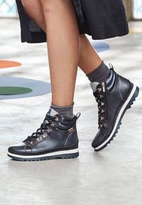 Baskets montantes noires en cuir lisse, avec œillets en métal, semelle blanche contrastante et détail de fermeture éclair sur le côté, accompagnées de chaussettes remontant jusqu'aux chevilles grises.