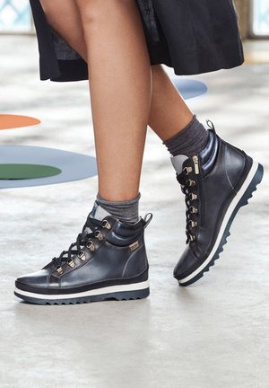 Baskets montantes noires en cuir lisse, avec œillets en métal, semelle blanche contrastante et détail de fermeture éclair sur le côté, accompagnées de chaussettes remontant jusqu'aux chevilles grises.