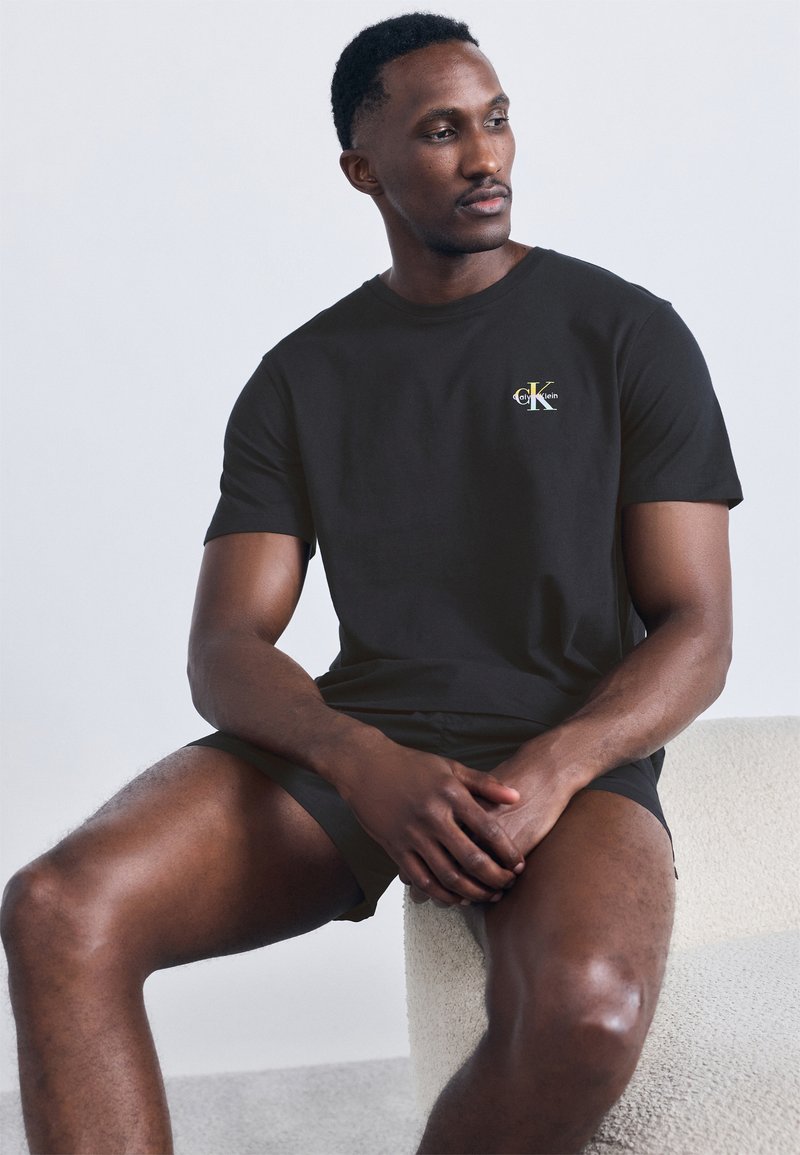 Homme assis sur une chaise blanche texturée portant un t-shirt noir Calvin Klein et un short assorti, regardant vers sa droite.
