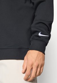 Svart atletisk hettegenser med myk tekstur, med ribbestrikkede mansjetter og en hvit Nike-logo på venstre erme. Nærbilde av hånden ved mansjetten.