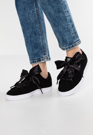 Person, die schwarze Wildledersneaker mit breiten Satinband-Schnürsenkeln und blaue Jeans aus Denim trägt und auf einem weißen Boden steht.