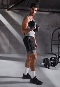 Haltere preto segurado com uma mão, vestindo uma camisola cinza, calções pretos com detalhes cinzentos e sapatos atléticos pretos com meias brancas.