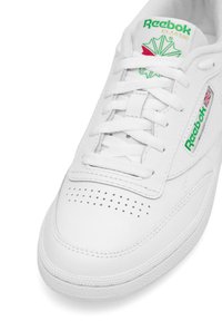 Valged tennisajad, mis on valmistatud siledast sünteetilisest nahast, perforeeritud nina detailsusega, Reeboki roheline ja punane logo ning klassikalise paeltega disain.