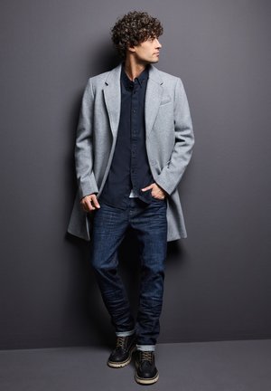 Abrigo de lana gris sobre una camisa oscura de botones, combinado con unos vaqueros de denim azul y botas negras con cordones. El fondo es una pared gris sólida.