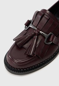 Burgundské kožené loafery s fringingovými strapcami a čiernymi kovovými doplnkami. Majú zaoblenú špičku a pletenú čiernu podrážku. Celkový hladký povrch.