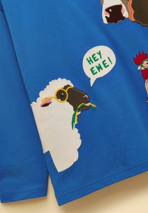 Mouton blanc au visage noir portant des lunettes jaunes, mâchant de l'herbe verte, bulle de dialogue avec le texte "HEY EWE !" sur un fond en tissu bleu.