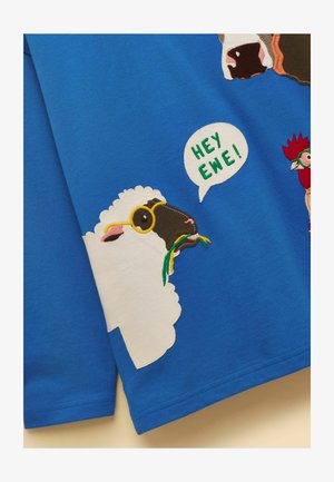 Valkoinen lammas, musta pää ja keltaiset silmälasit, pureskelee vihreää ruohoa, puhekupla, jossa teksti "HEY EWE!" sinisellä kangastaustalla.