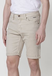 Man som bär beige denimshorts med upprullade sidor och en vit t-shirt instoppad, står mot en enfärgad bakgrund.