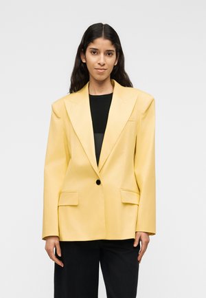 Vrouw met lang donker haar draagt een oversized gele blazer over een zwart shirt en zwarte broek, staand tegen een grijze achtergrond.