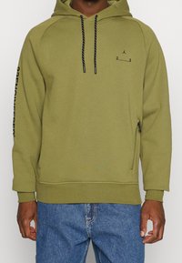 Hombre con sudadera con capucha verde oliva con cordones negros, pequeño logo negro y números en el pecho, texto negro en la manga izquierda y bolsillo con cremallera en el costado.